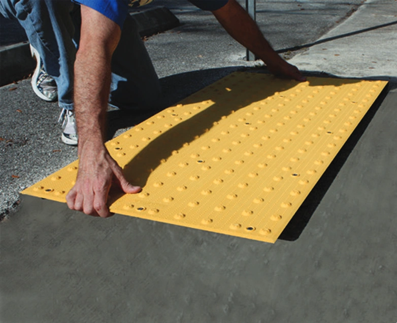Construction worker in orange shirt kneeling to install yellow tactile paving with raised domes, aligning surface using a spirit level on fresh concrete sidewalk.
