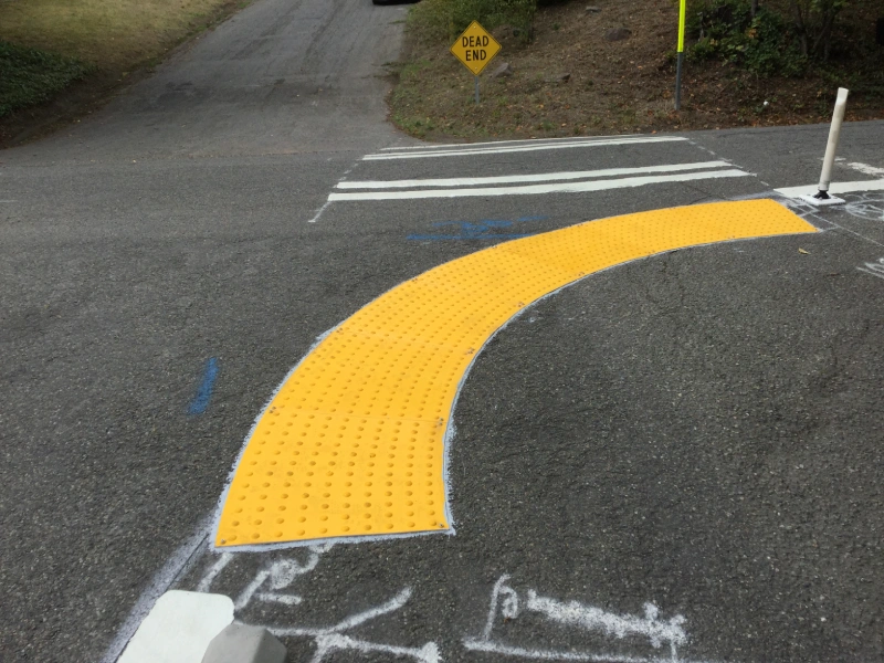 Curved yellow tactile paving with raised domes installed at street corner, guiding pedestrians toward crosswalk for accessibility and safety.