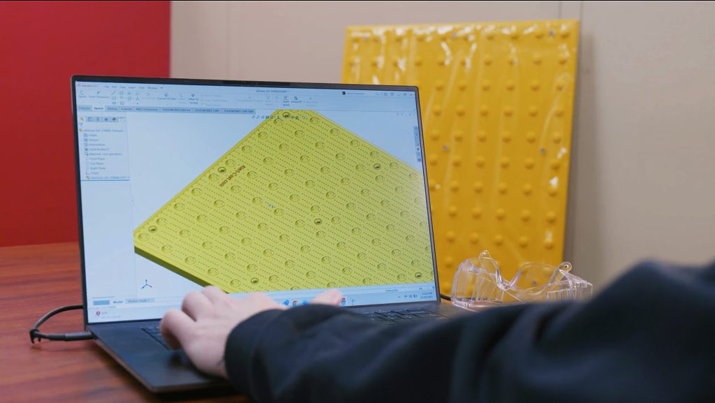 Laptop displaying 3D CAD model of yellow tactile paving tile with raised bumps, alongside physical prototype leaning against wall in industrial workspace.