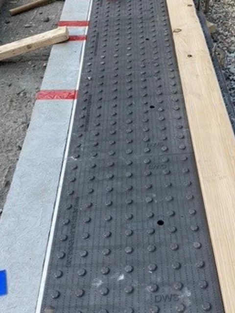 Black tactile paving with raised domes installed on sidewalk under construction, bordered by concrete and wooden plank with red tape markings.