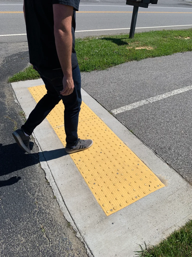 Person walking on sidewalk and stepping onto yellow tactile paving with raised domes at curb ramp, designed for accessibility and safe street crossing.