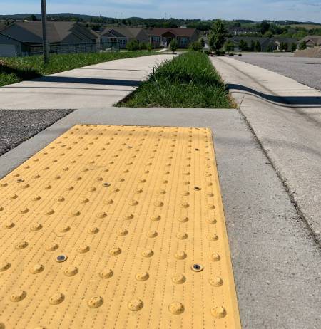Smoothly sloped curb ramp with yellow tactile paving featuring raised domes, providing accessibility and safety for pedestrians at residential sidewalk crossing.